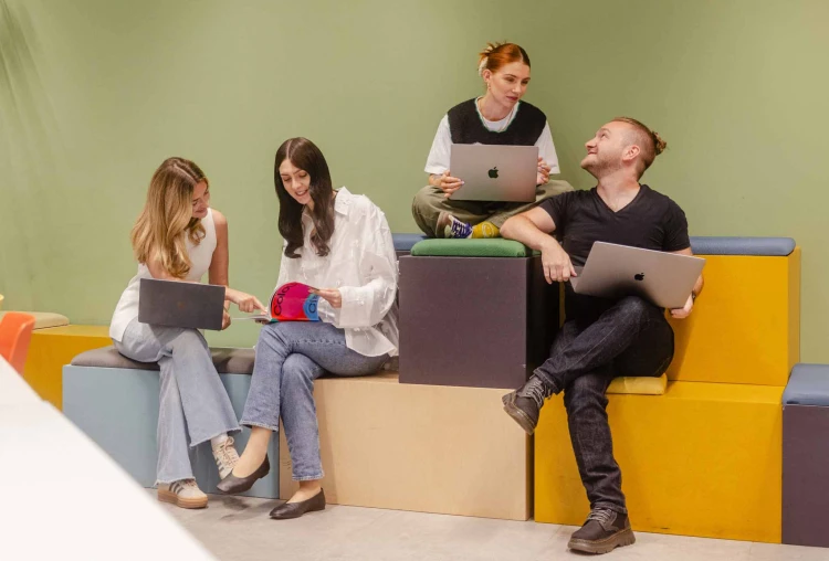 Three women one man sat on stools talking smiling with laptops.